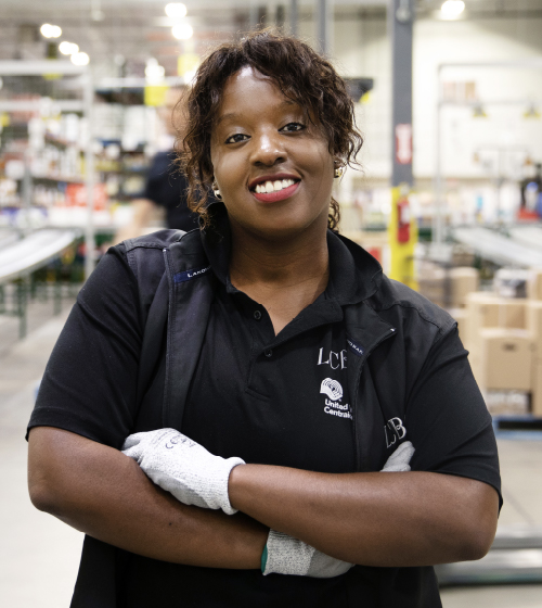 A female LCBO warehouse employee with a bright smile, wearing white work gloves standing in an LCBO Warehouse. 