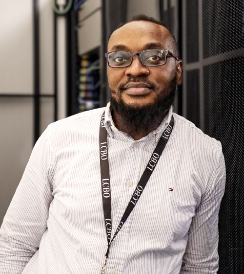 A male employee with glasses and a beard wearing a white shirt standing in front of the technology office at LCBO headquarters.